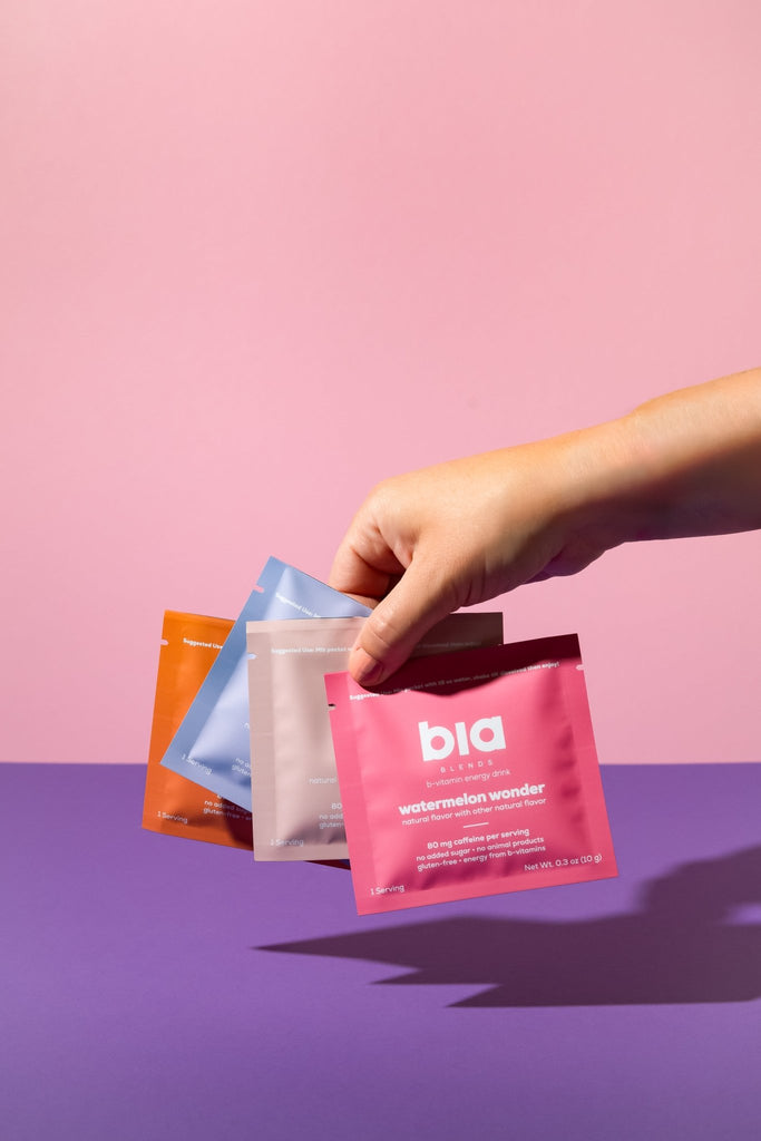 Hand holding four healthy energy drink sample packet with 'bia' branding against a pink and purple gradient background.